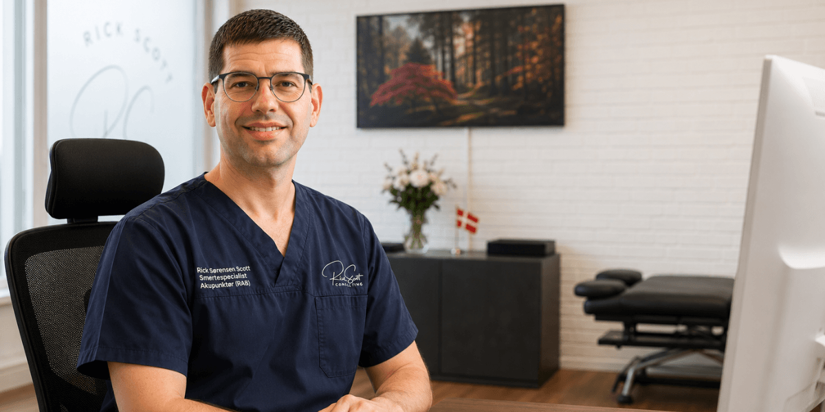 A smiling professional man in scrubs sits at an office desk with medical equipment.