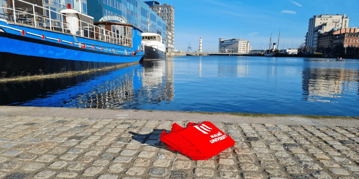 Red Malmö University tote bag displayed beside a waterfront canal in Malmö, Sweden.
