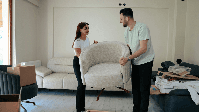 This image shows a man and a woman in the middle of a move or furniture arrangement. They are both smiling and appear to be lifting a modern, beige armchair together. In the background, a spacious living room is being set up, with a couple of couches and cardboard boxes scattered around. The atmosphere feels light-hearted and collaborative, suggesting they are in the process of settling into a new space.
