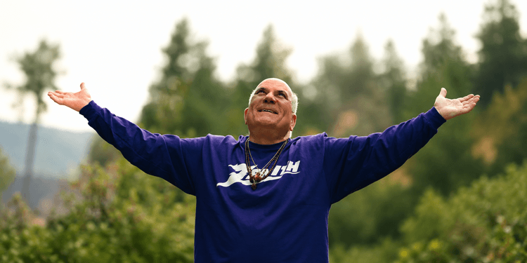 Man in a purple shirt with arms outstretched, smiling against a forest backdrop