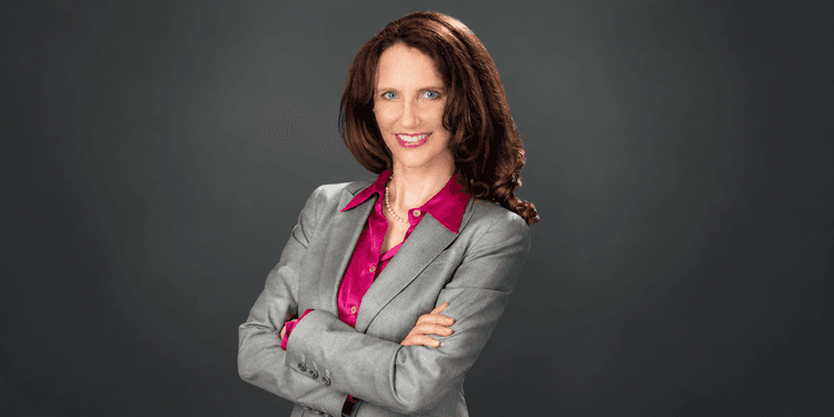 Woman in a gray suit with crossed arms and a pink blouse, smiling confidently