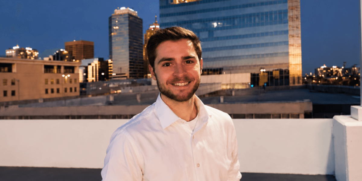 Smiling young man in a white shirt stands against a city skyline at dusk