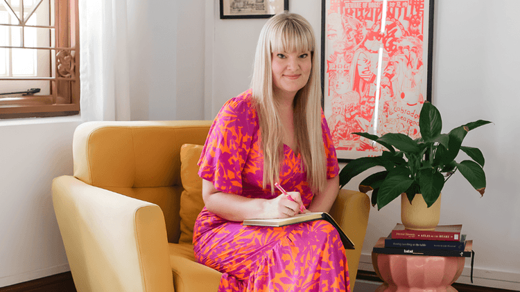 Nikki Smit sits on a yellow chair, writing in a notebook with a plant nearby.