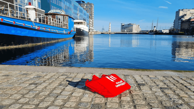 Red Malmö University tote bag displayed beside a waterfront canal in Malmö, Sweden.