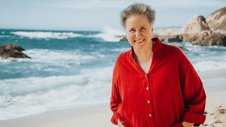 A smiling woman in a red shirt standing on a beach, with the ocean waves and rocky cliffs in the background, creating a serene and natural setting.