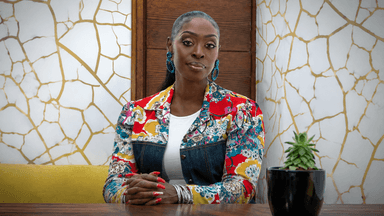 Dilys Sillah seated at a table wearing a colorful floral jacket in a modern interior.