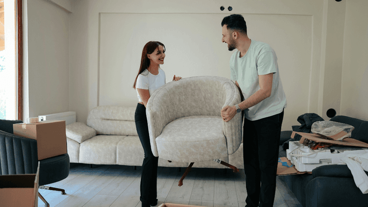 This image shows a man and a woman in the middle of a move or furniture arrangement. They are both smiling and appear to be lifting a modern, beige armchair together. In the background, a spacious living room is being set up, with a couple of couches and cardboard boxes scattered around. The atmosphere feels light-hearted and collaborative, suggesting they are in the process of settling into a new space.
