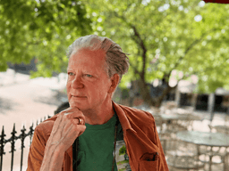 Man with gray hair wearing a green shirt and brown jacket outdoors, pondering thoughtfully