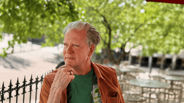 Man with gray hair wearing a green shirt and brown jacket outdoors, pondering thoughtfully