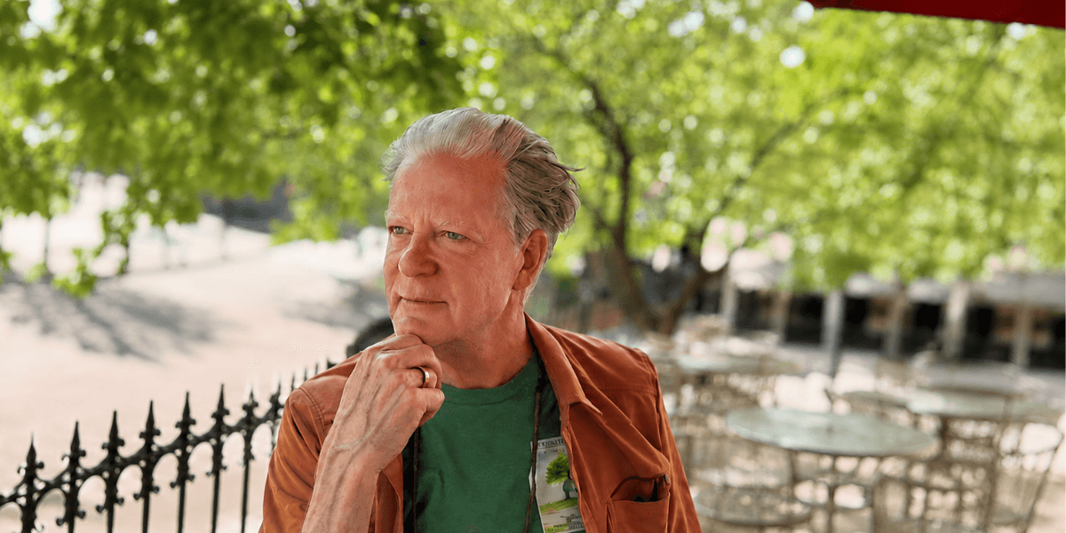 Man with gray hair wearing a green shirt and brown jacket outdoors, pondering thoughtfully