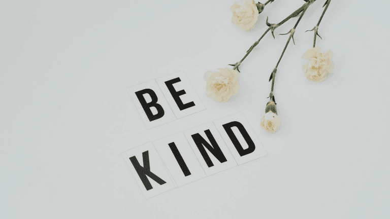 Flat lay of white flowers and letter tiles spelling "BE KIND" on a light background