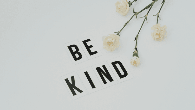 Flat lay of white flowers and letter tiles spelling "BE KIND" on a light background