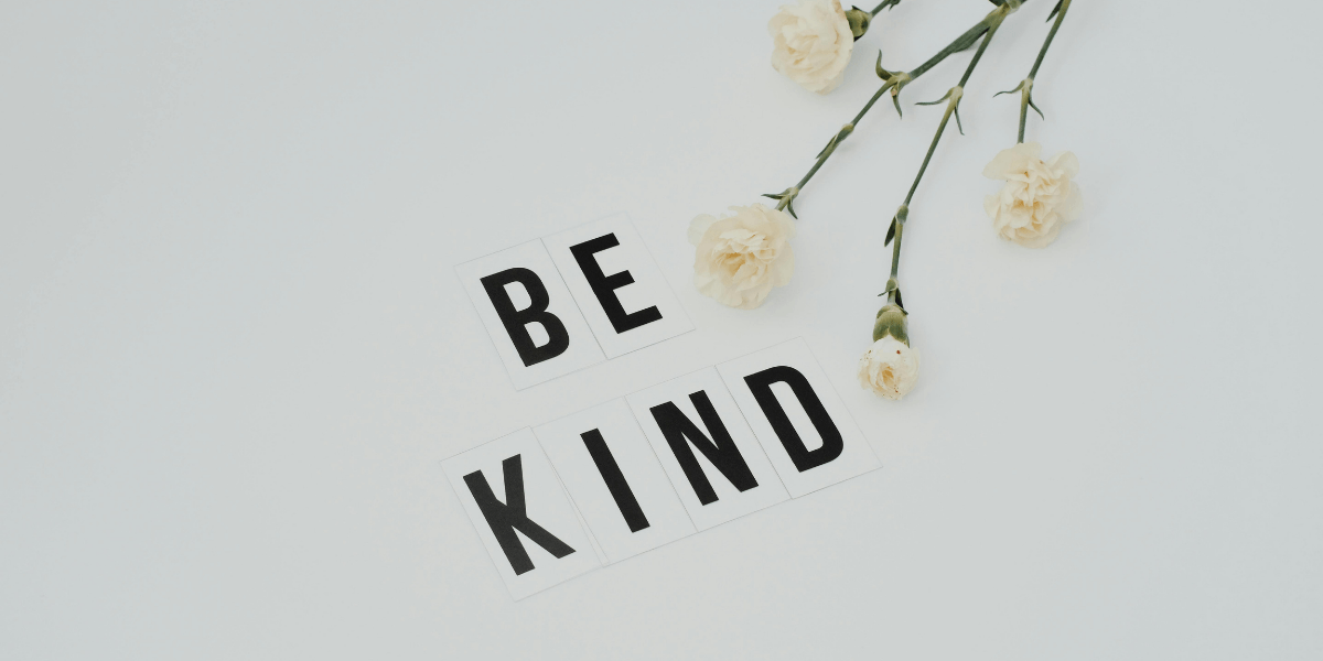 Flat lay of white flowers and letter tiles spelling "BE KIND" on a light background