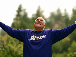 Man in a purple shirt with arms outstretched, smiling against a forest backdrop