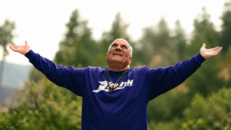 Man in a purple shirt with arms outstretched, smiling against a forest backdrop
