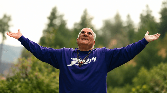 Man in a purple shirt with arms outstretched, smiling against a forest backdrop