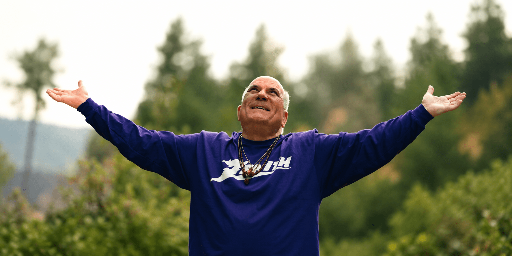 Man in a purple shirt with arms outstretched, smiling against a forest backdrop