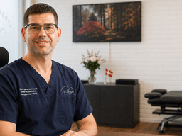 A smiling professional man in scrubs sits at an office desk with medical equipment.