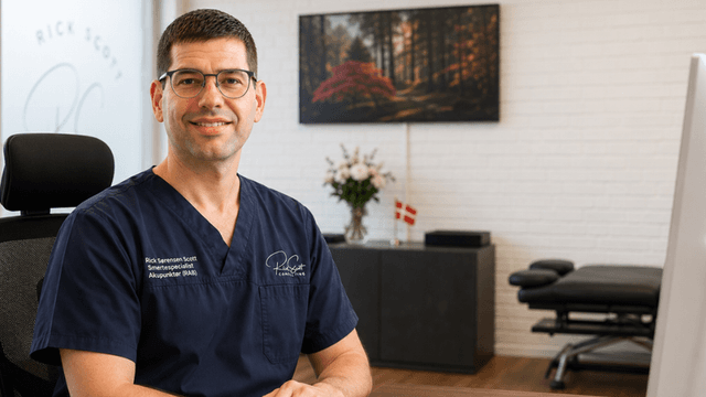A smiling professional man in scrubs sits at an office desk with medical equipment.