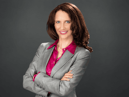 Woman in a gray suit with crossed arms and a pink blouse, smiling confidently