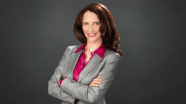 Woman in a gray suit with crossed arms and a pink blouse, smiling confidently