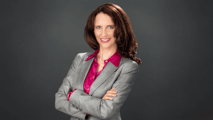 Woman in a gray suit with crossed arms and a pink blouse, smiling confidently