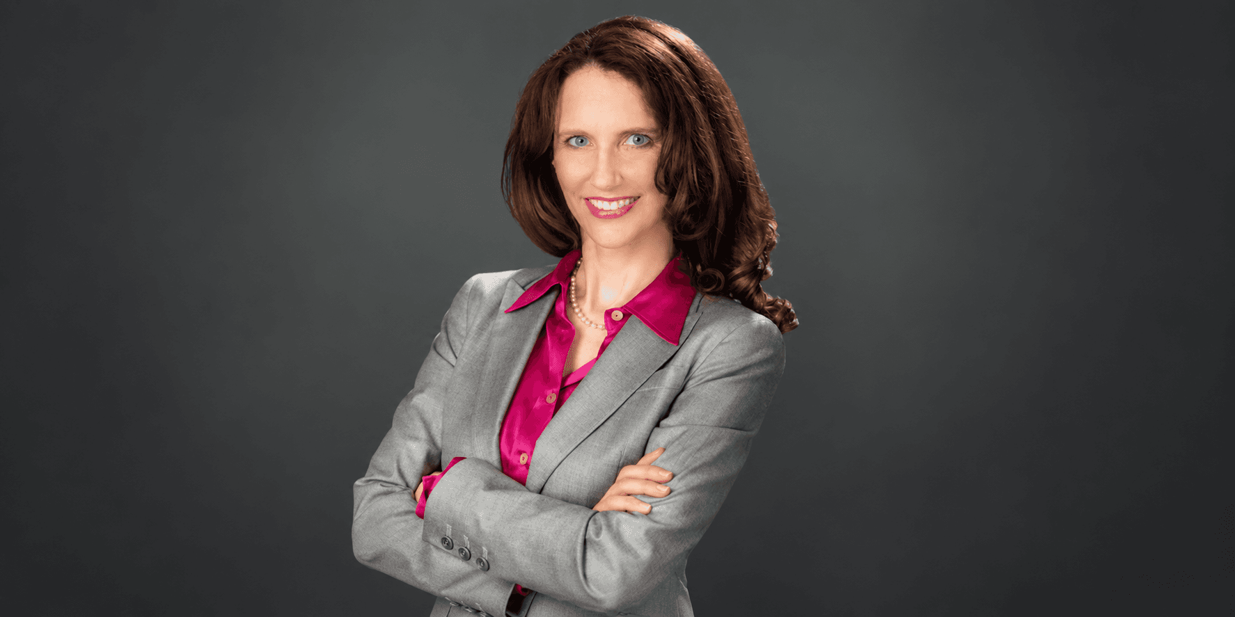 Woman in a gray suit with crossed arms and a pink blouse, smiling confidently
