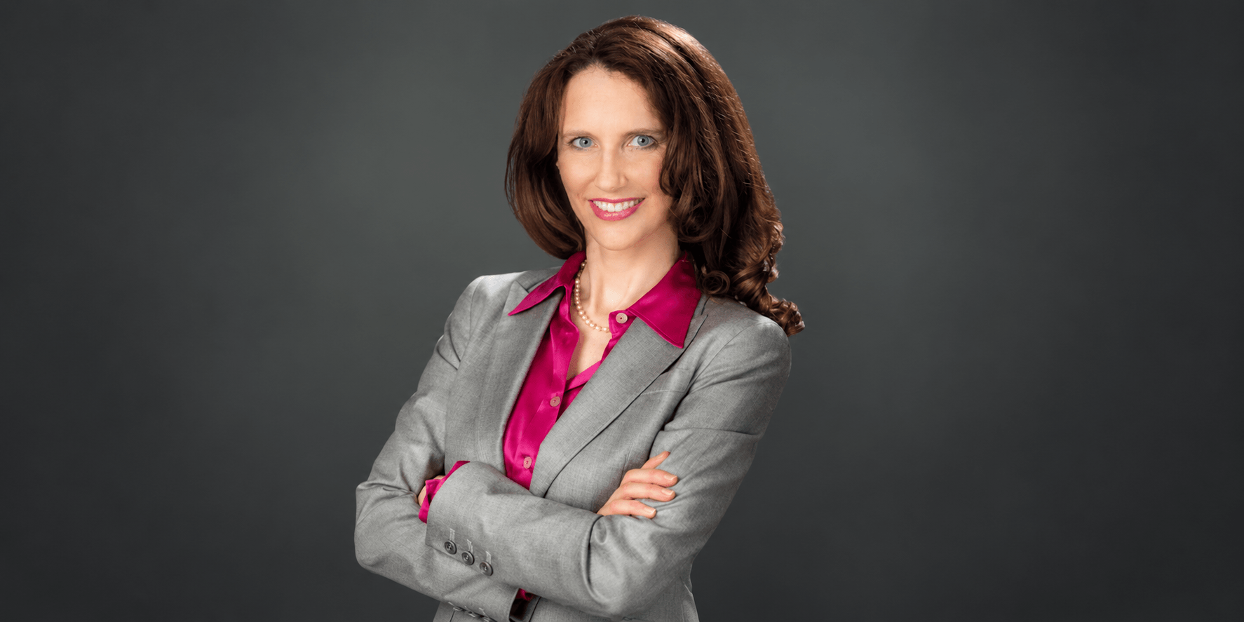 Woman in a gray suit with crossed arms and a pink blouse, smiling confidently