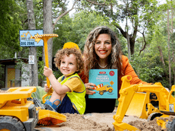 A smiling woman and a child play in a sandpit with toy construction vehicles.
