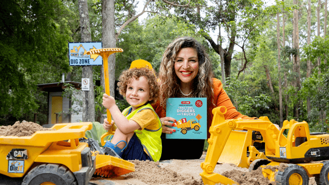 A smiling woman and a child play in a sandpit with toy construction vehicles.