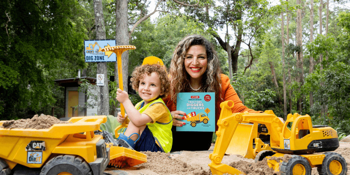 A smiling woman and a child play in a sandpit with toy construction vehicles.