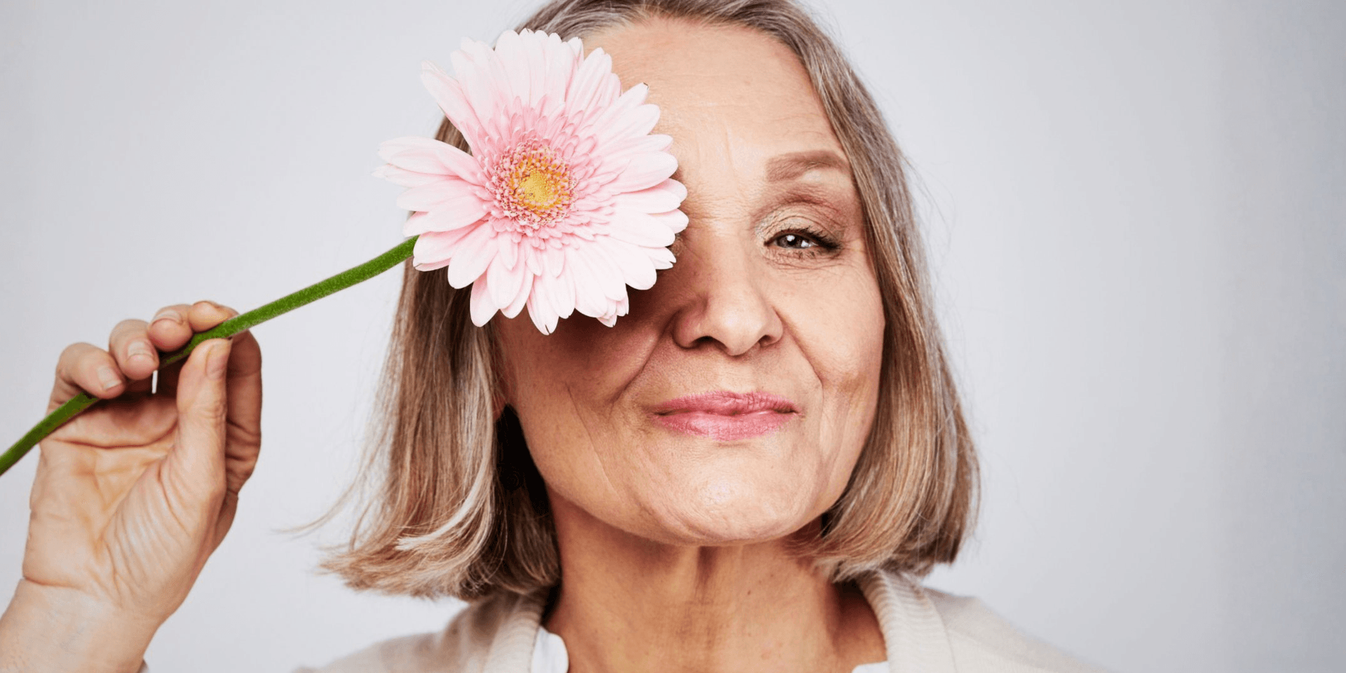 Smiling older woman holding a pink flower next to her face against a plain background