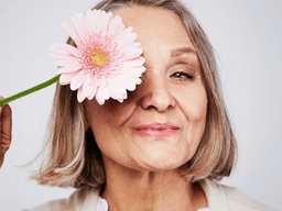 Smiling older woman holding a pink flower next to her face against a plain background
