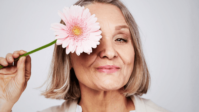 Smiling older woman holding a pink flower next to her face against a plain background