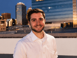 Smiling young man in a white shirt stands against a city skyline at dusk
