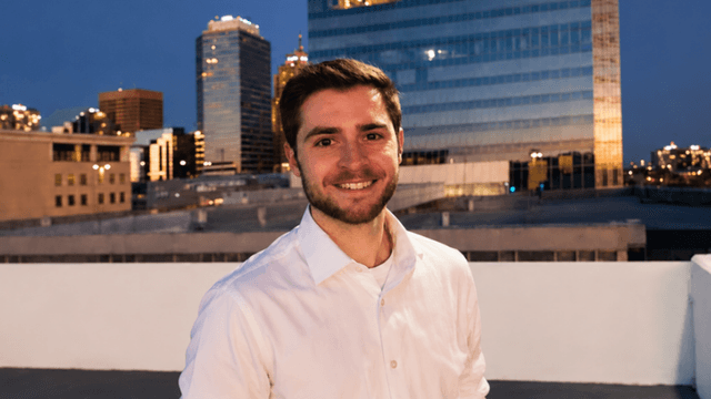 Smiling young man in a white shirt stands against a city skyline at dusk
