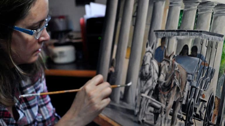 The image shows an artist painting a horse-drawn carriage with a focus on the details of the artwork being created.