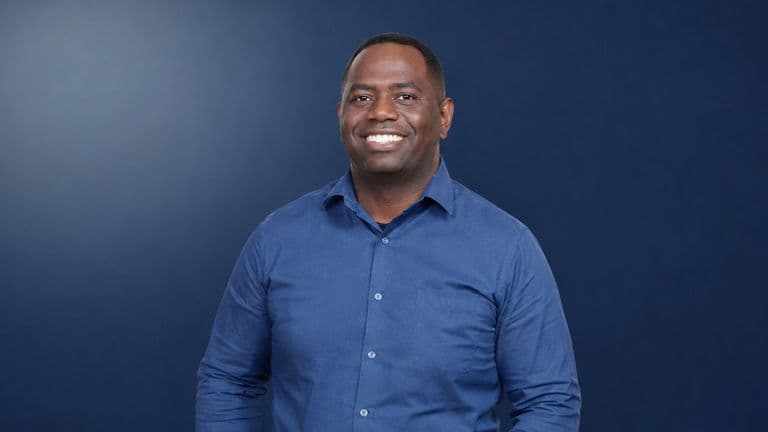 Smiling man in blue button-down shirt against dark blue background, accompanying article about podcast host fostering authentic conversations.