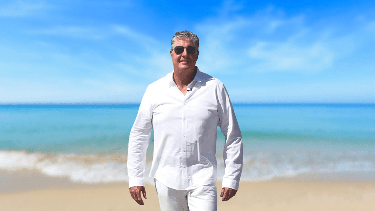 Kevin L. McMullen walks confidently along a beach, dressed in a white shirt and sunglasses, with a serene ocean view in the background.
