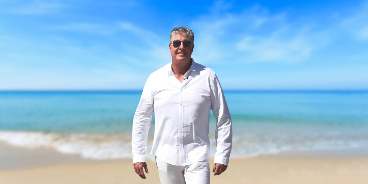 Kevin L. McMullen walks confidently along a beach, dressed in a white shirt and sunglasses, with a serene ocean view in the background.