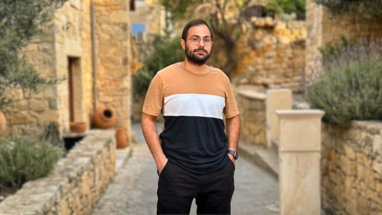 A man with glasses stands in front of a textured stone wall, embodying a thoughtful presence in a poetic setting.
