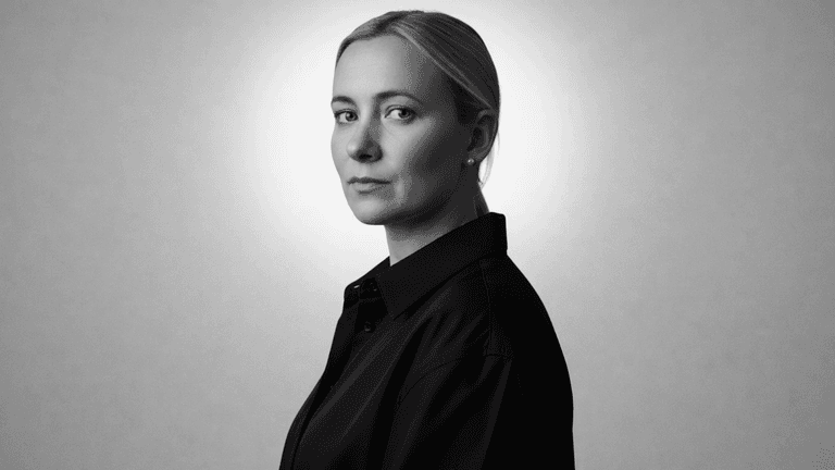Black-and-white portrait of woman in dark shirt, looking over shoulder against plain background.