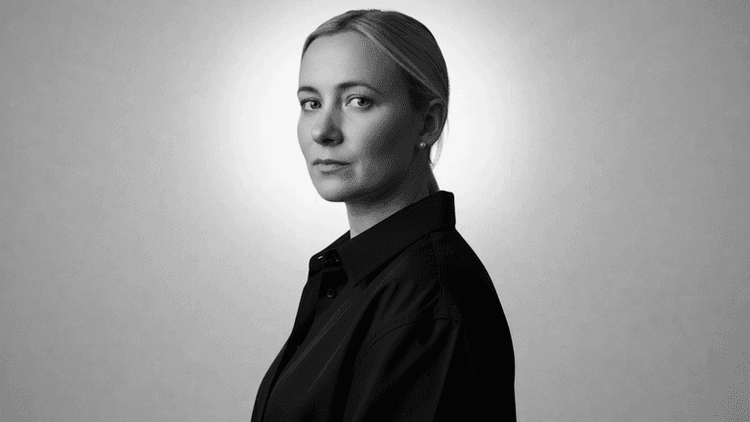 Black-and-white portrait of woman in dark shirt, looking over shoulder against plain background.