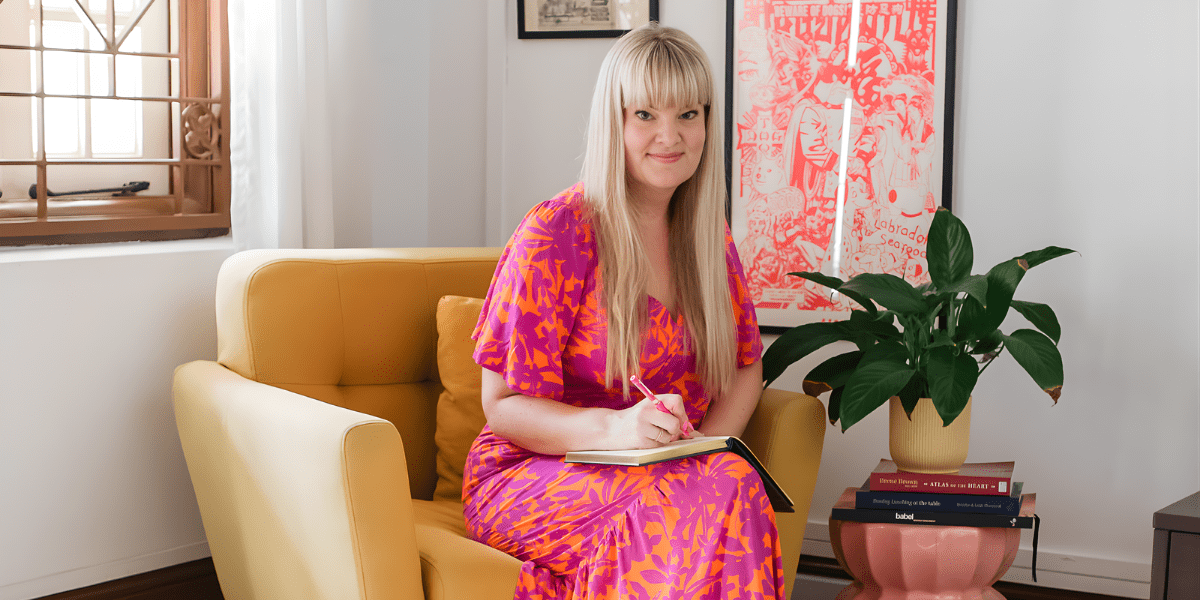 Nikki Smit sits on a yellow chair, writing in a notebook with a plant nearby.