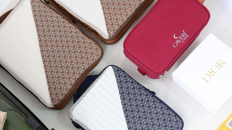 A collection of travel bags on a table, featuring brown and navy patterned cases, a red Caleri pouch, and a white Dior box. The scene conveys organization.