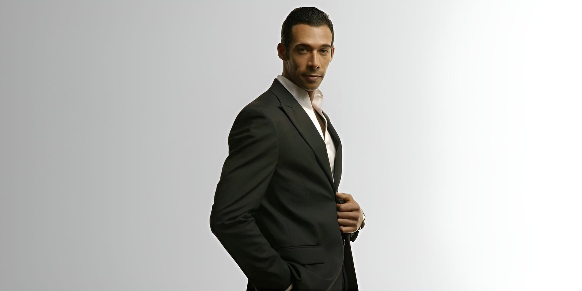 Man in dark suit adjusting jacket, looking confidently at camera against plain background.