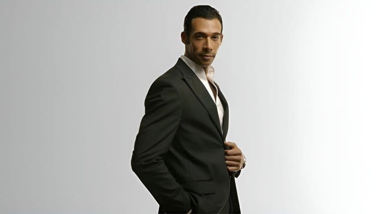 Man in dark suit adjusting jacket, looking confidently at camera against plain background.