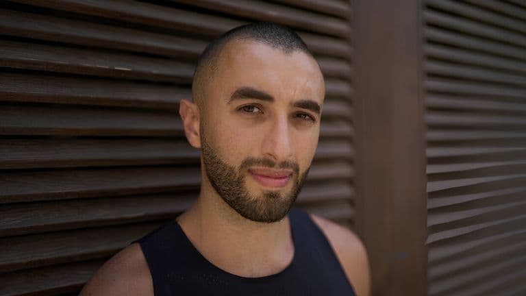 A man with a shaved head and beard gazes calmly at the camera, standing against a textured wooden slat wall. He wears a black tank top, creating a serene tone.