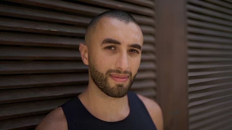 A man with a shaved head and beard gazes calmly at the camera, standing against a textured wooden slat wall. He wears a black tank top, creating a serene tone.