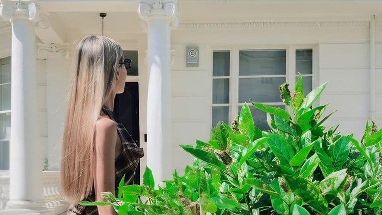 Woman with long blonde hair and sunglasses beside bright green plants outside white-columned house.